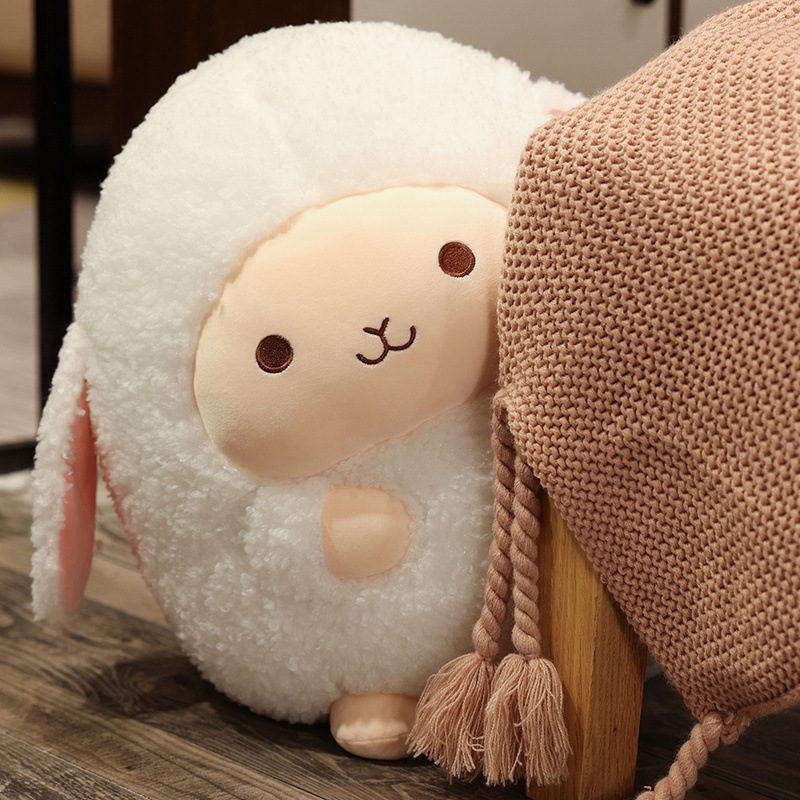 Round sheep plushie with peach face and pink ears sitting beside a textured blanket.