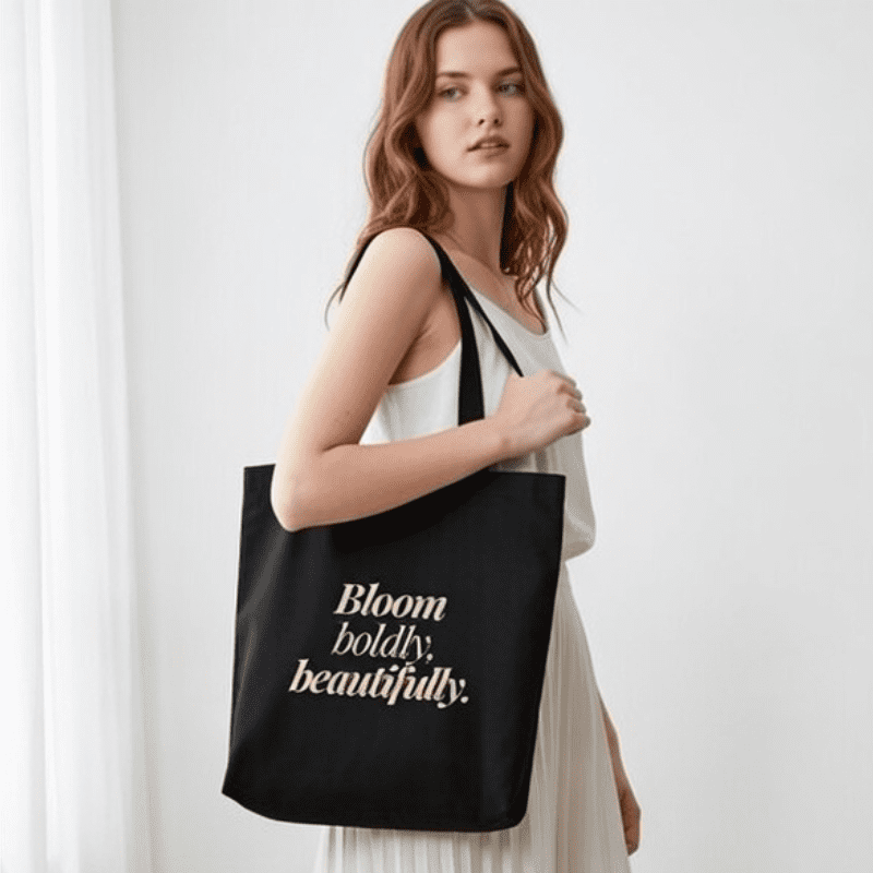 bloom_boldly_beautifully_tote_bag_quotequest365 Model carrying a Quote Quest 365 black canvas tote bag featuring the inspirational quote 'Bloom Boldly, Beautifully.' in gold elegant font, perfect for #onthegoessentials.