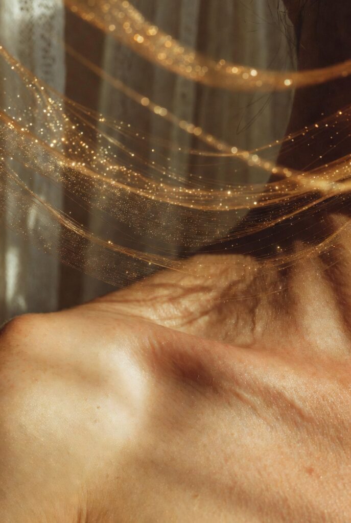 Close-up of a person’s shoulder and neck with delicate, shimmering golden threads and sparkling particles draped across warm-toned skin, creating an ethereal and luminous artistic effect.