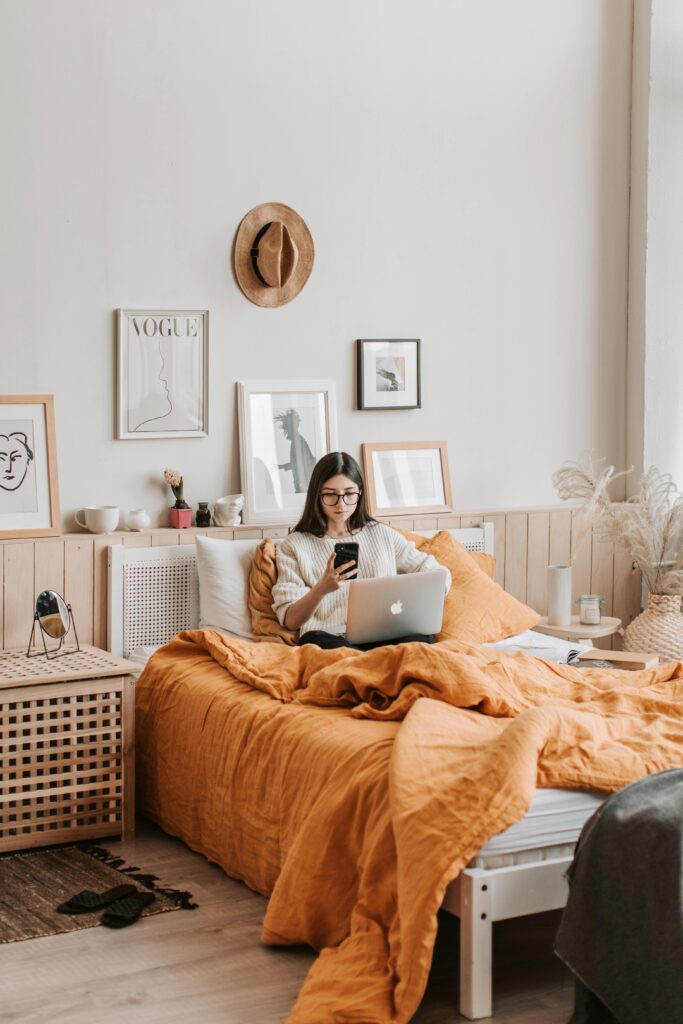 A cozy bedroom scene with a woman wearing glasses sitting on an orange-bedded bed, working on her laptop while holding her phone, surrounded by framed art and warm decor.