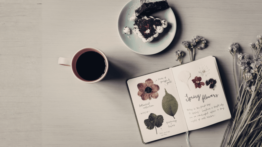 Top-down view of black coffee in pink-rimmed mug, chocolate cake slice, open notebook with pressed flowers and handwritten notes, dried flowers on light wooden table